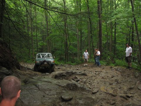 1995 Ford C-350 (Centurion Conversions) - Off-road vehicle tackles a challenging muddy path in a dense forest environment.