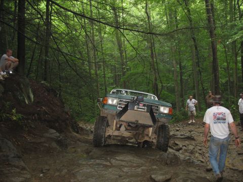 1995 Ford C-350 (Centurion Conversions) - An off-road vehicle tackles a challenging rocky trail surrounded by lush green trees.