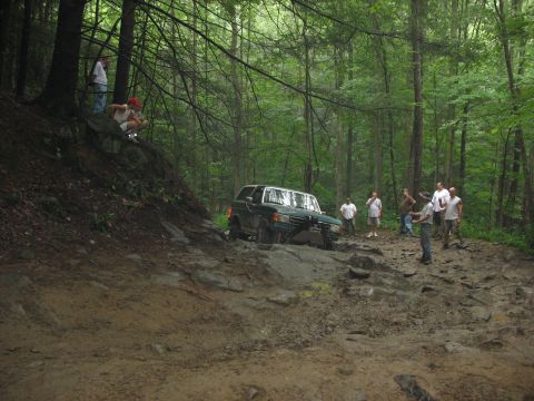 1995 Ford C-350 (Centurion Conversions) - A green Ford Bronco navigates a challenging off-road trail in a wooded area.
