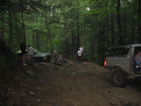 1995 Ford C-350 (Centurion Conversions) - People gather around two off-road vehicles stuck in the mud on a wooded trail.