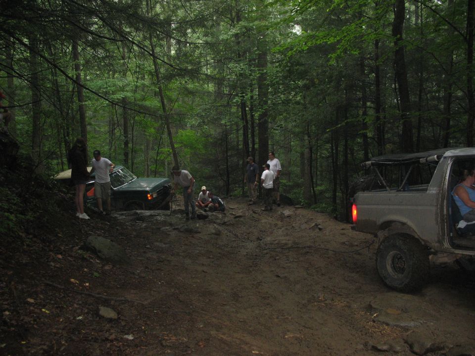 Two off-road vehicles stuck in mud on a forest trail with people around