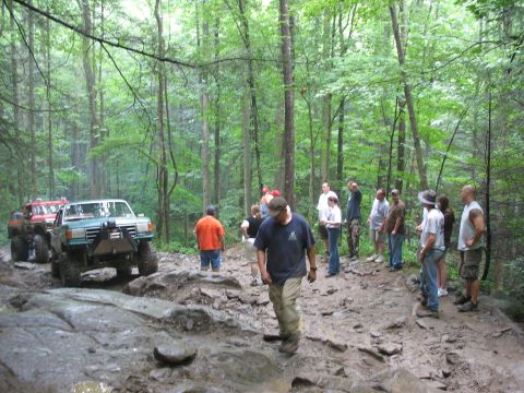 1995 Ford C-350 (Centurion Conversions) - Two trucks navigate a challenging muddy and rocky trail through a dense forest with onlookers.