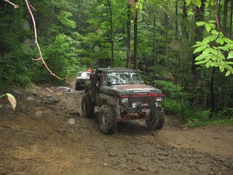 1995 Ford C-350 (Centurion Conversions) - A heavily modified truck navigates a challenging muddy trail in a forest, with another vehicle following closely.