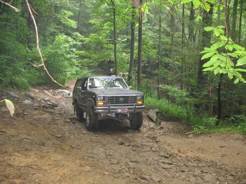 1995 Ford C-350 (Centurion Conversions) - Dark SUV navigates a challenging muddy and rocky trail in a dense forest setting.