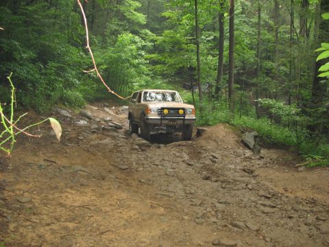 1995 Ford C-350 (Centurion Conversions) - A tan Ford Bronco navigates a challenging, wet, and rocky off-road trail surrounded by dense green forest.