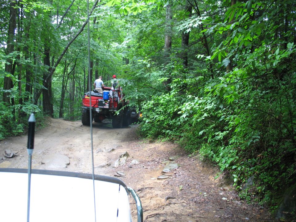 Two off-road vehicles driving on a muddy, wooded trail. One red truck is ahead, and the shot is from inside another vehicle