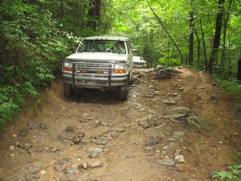 1995 Ford C-350 (Centurion Conversions) - Two trucks navigate a challenging, rocky, and muddy path through a dense forest.