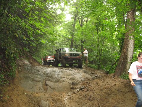 1995 Ford C-350 (Centurion Conversions) - Several off-road vehicles are seen traversing a challenging, wet, and rocky trail through a dense forest.
