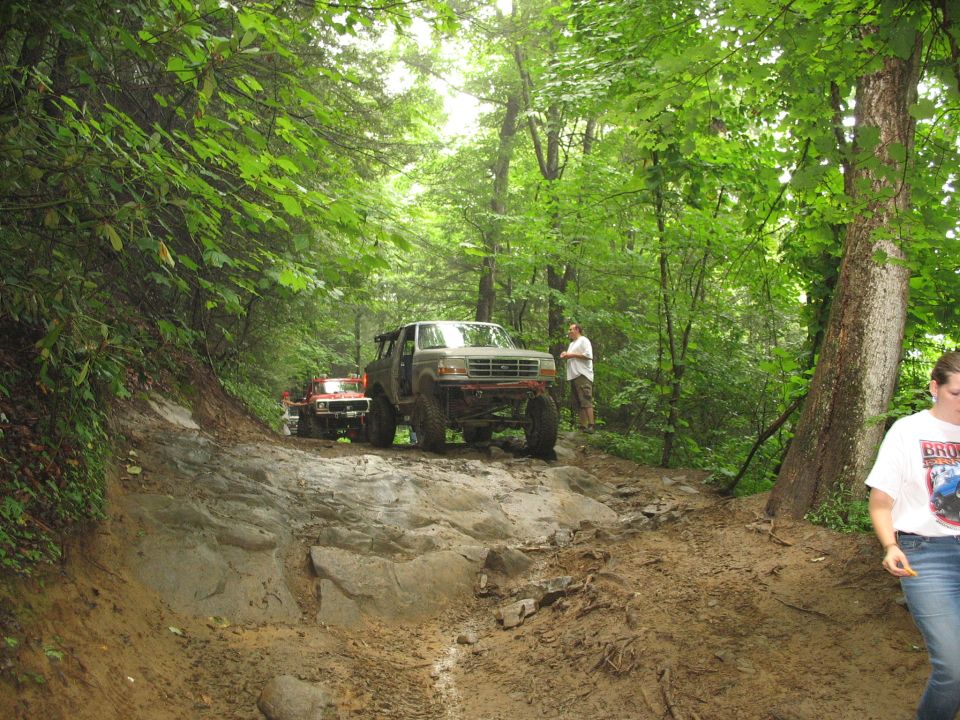 Modified Ford Bronco or F-Series truck navigating a rocky, muddy trail in a forest. Another red off-road vehicle is behind it