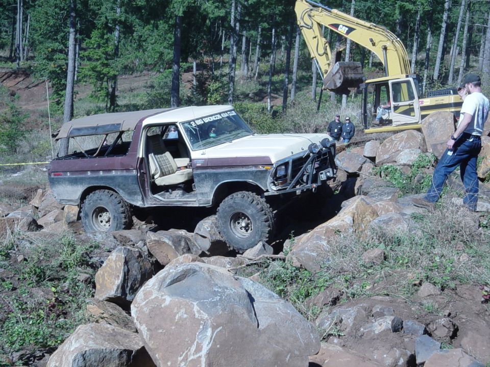 Tan and maroon Ford Bronco winched over rocks with a CAT excavator alongside