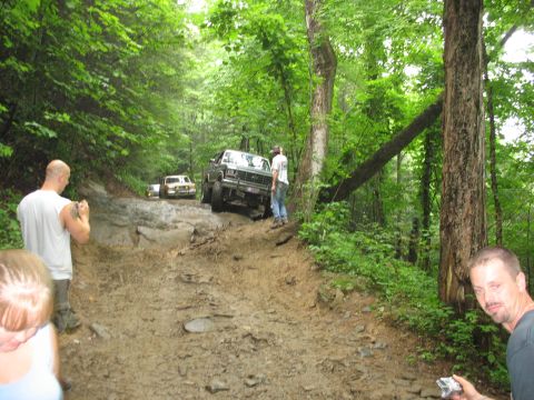 1995 Ford C-350 (Centurion Conversions) - Several off-road vehicles navigate a challenging, muddy, and rocky trail through a dense forest.
