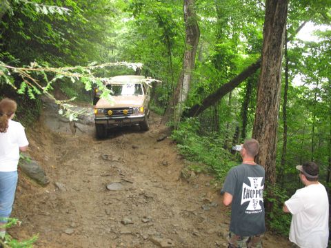 1995 Ford C-350 (Centurion Conversions) - An off-road vehicle navigates a challenging, muddy path through a dense forest.