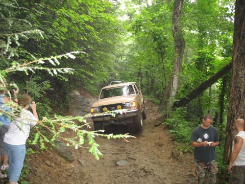 1995 Ford C-350 (Centurion Conversions) - A tan SUV navigates a challenging muddy path through a dense forest, observed by onlookers.