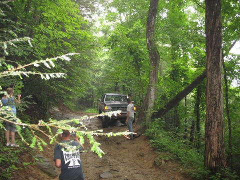 1995 Ford C-350 (Centurion Conversions) - A dark pickup truck navigates a muddy trail through a dense forest, with onlookers observing.