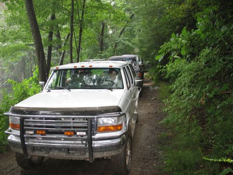 1995 Ford C-350 (Centurion Conversions) - Several white SUVs, including a 1995 Ford C-350 (Centurion Conversions), drive down a wooded dirt road.