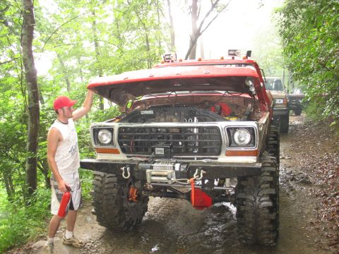 1995 Ford C-350 (Centurion Conversions) - A man inspects a red Ford truck with its hood open, parked on a muddy trail behind other vehicles.
