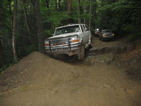 1995 Ford C-350 (Centurion Conversions) - Silver SUV navigates a challenging muddy and rocky path through a dense forest with other vehicles following.