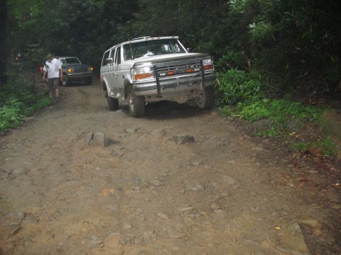 1995 Ford C-350 (Centurion Conversions) - Ford Bronco driving on a challenging, rocky dirt path through a wooded area.