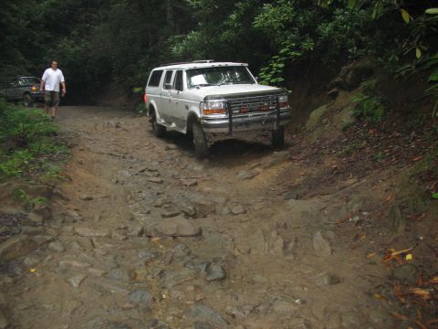 1995 Ford C-350 (Centurion Conversions) - A white SUV navigates a challenging muddy and rocky path through a dense forest.