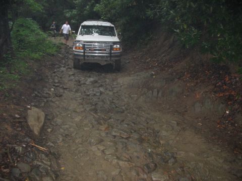 1995 Ford C-350 (Centurion Conversions) - A white Ford F-Series truck navigates a challenging, rocky off-road path surrounded by dense foliage.