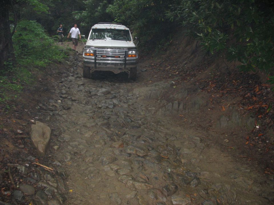 White Ford F-Series truck with aftermarket grille guard driving on a rocky, muddy trail