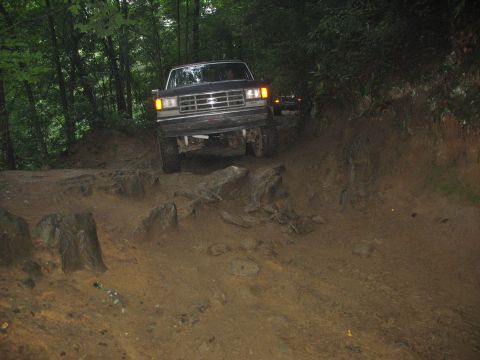1995 Ford C-350 (Centurion Conversions) - A Ford truck is shown driving on a rough, dirt path surrounded by trees. Another vehicle is visible behind it.