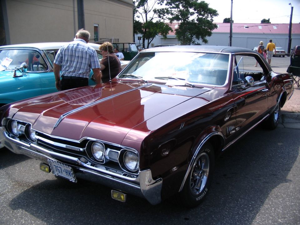 Maroon 1967 Oldsmobile Cutlass Supreme parked next to a teal car. People stand between them