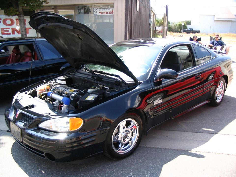Black Pontiac Grand Am coupe, hood open showing engine. Another black car is parked to the left. Two people sit in chairs in the background