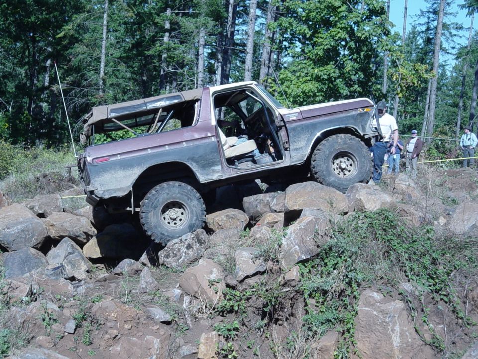 Maroon and tan Ford Bronco crawling rocks, doors open, spotters watching at right