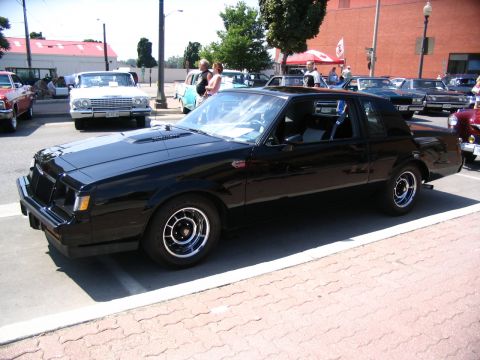 1993 Ford Taurus - A black coupe with a distinctive grille and hood scoop is parked on a brick-lined street, with other vehicles and pedestrians around.