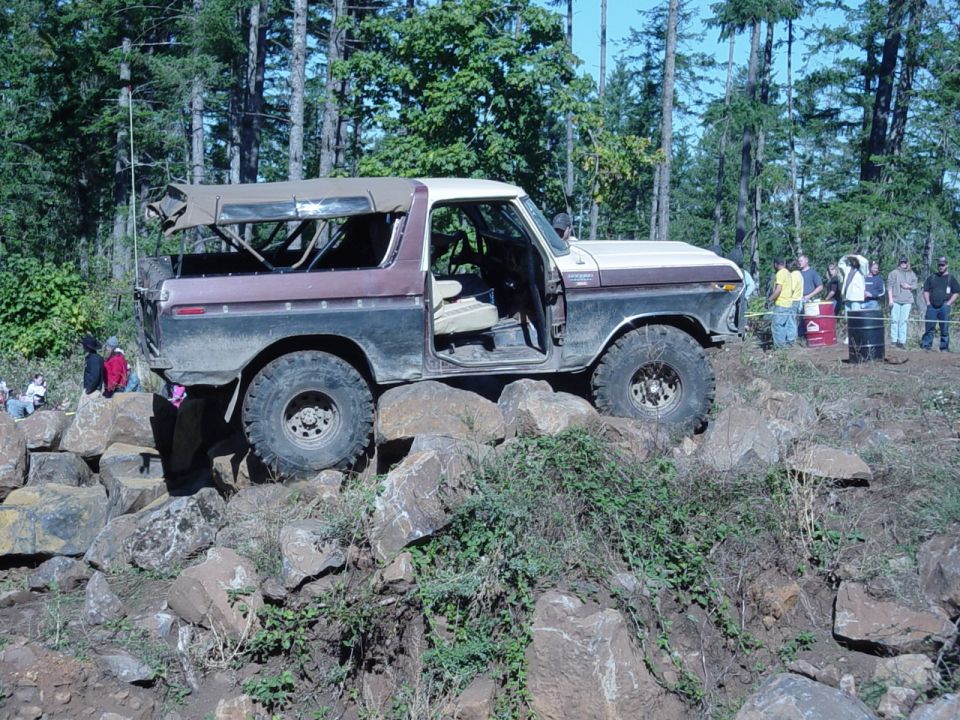 Tan and maroon Ford Bronco on a rocky ridge, top folded, crowd of spectators right