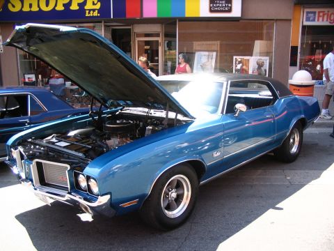 1993 Ford Taurus - A blue coupe with its hood open is displayed at a car show, with other vehicles and a storefront in the background.