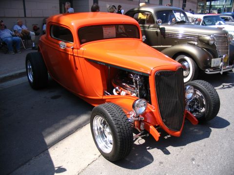 1993 Ford Taurus - An orange hot rod coupe is parked next to a classic car at a show.