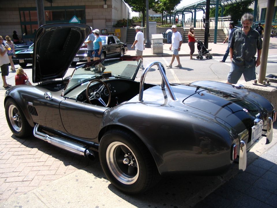 Dark grey convertible sports car with hood open, side pipes, and roll bar. Another car is visible behind it