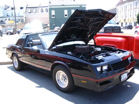 1993 Ford Taurus - A black Chevrolet Monte Carlo with its hood open, revealing the engine bay. The car is parked outdoors at a car show.