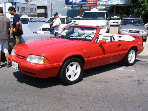 1993 Ford Taurus - A red convertible Mustang is parked on a street with other vehicles and people around.