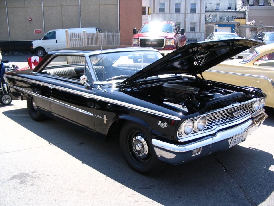 Black 1963 Ford Galaxie with hood open, parked next to a yellow car. A red GMC truck and white van are in the background