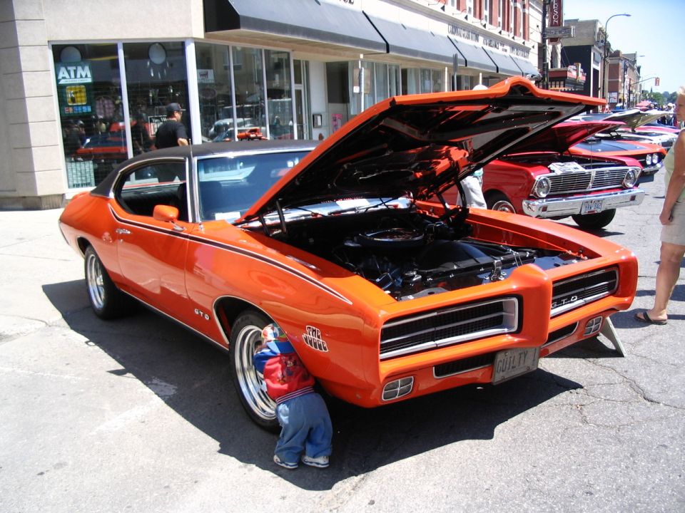 Orange 1969 Pontiac GTO with hood open, child looking at front wheel. Other cars in background