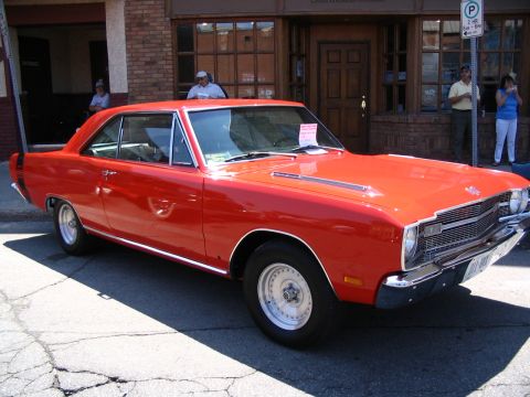 1993 Ford Taurus - A classic red Dodge Dart is displayed at a car show in front of a brick building.