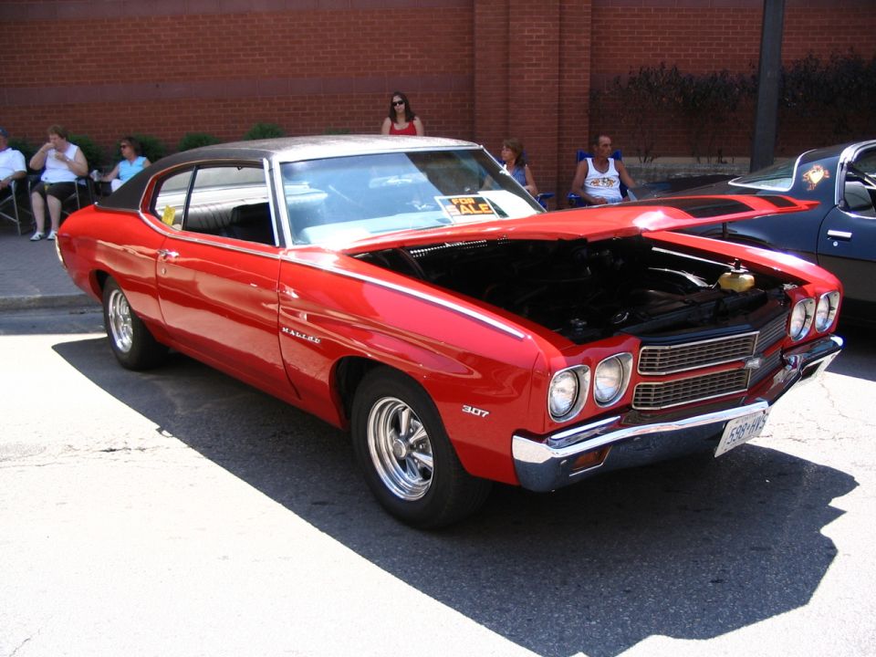 Red 1970s coupe with hood open, parked outside. Another car is visible to the right