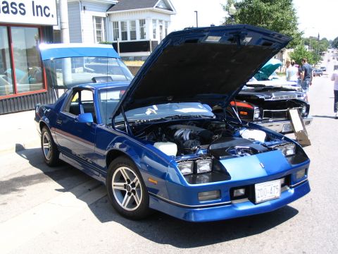 1993 Ford Taurus - A blue third-generation Chevrolet Camaro is displayed with its hood open at a car show. The vehicle is parked in front of a building with a…
