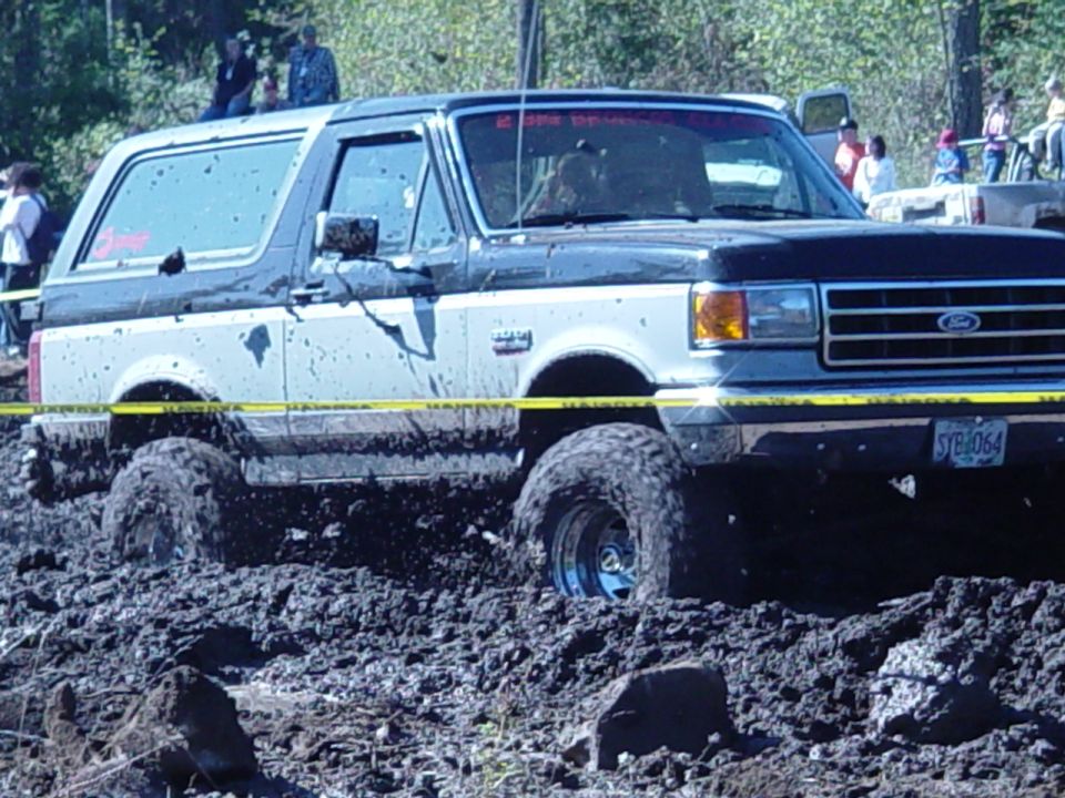 Silver and black Ford Bronco half-buried in a mud pit, spectators behind caution tape