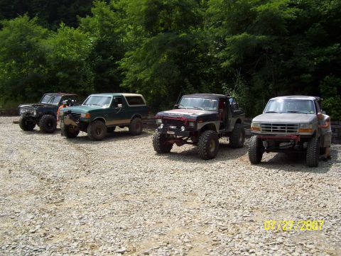 1978 Ford Bronco - Several off-road vehicles are parked in a lot with a wooded area behind them.