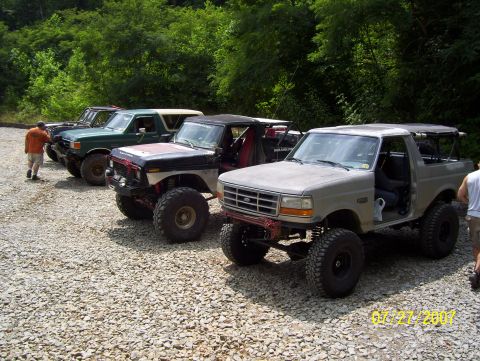 1978 Ford Bronco - Several modified Ford Broncos are lined up on a gravel lot with trees in the background.