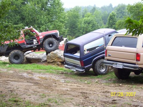 1978 Ford Bronco - Three modified off-road vehicles navigate a muddy terrain with trees in the background.