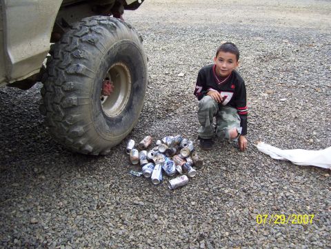 1978 Ford Bronco - A boy kneels by a pile of crushed cans next to the massive tire of an off-road vehicle.