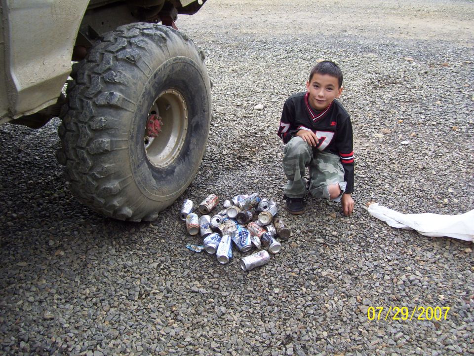 Large off-road tire and wheel of a light-colored vehicle, with a boy and crushed cans on gravel