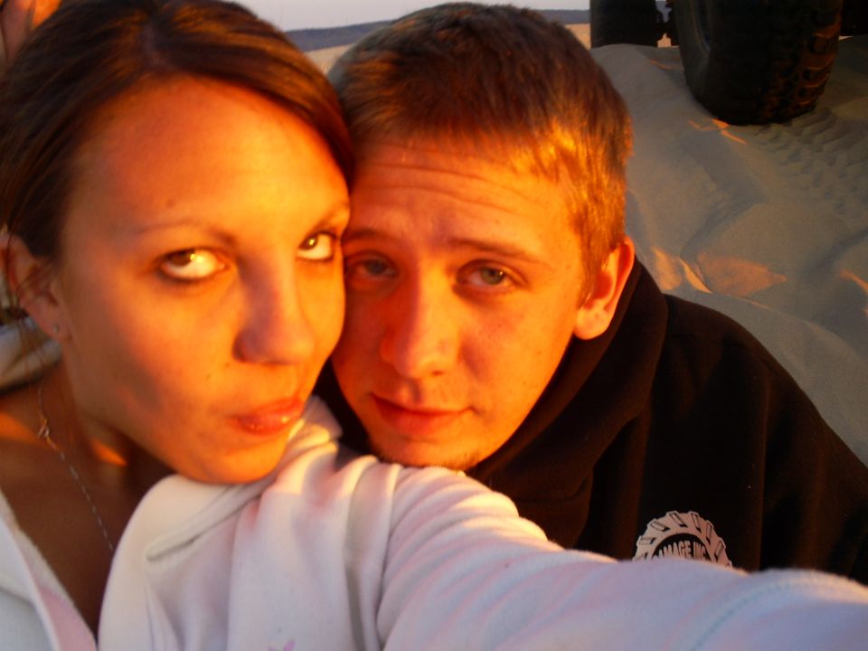 Couple posing for a selfie on sand dunes with a tire visible in the background