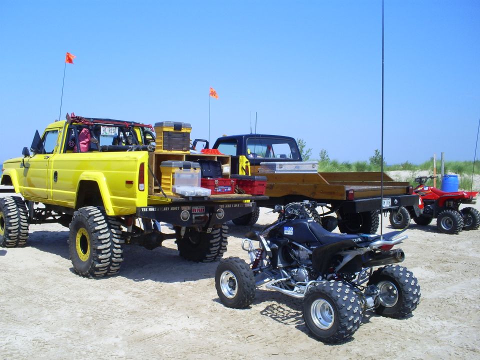 Yellow Jeep J-10 pickup truck with large tires, towing a flatbed trailer with ATVs on a sandy beach