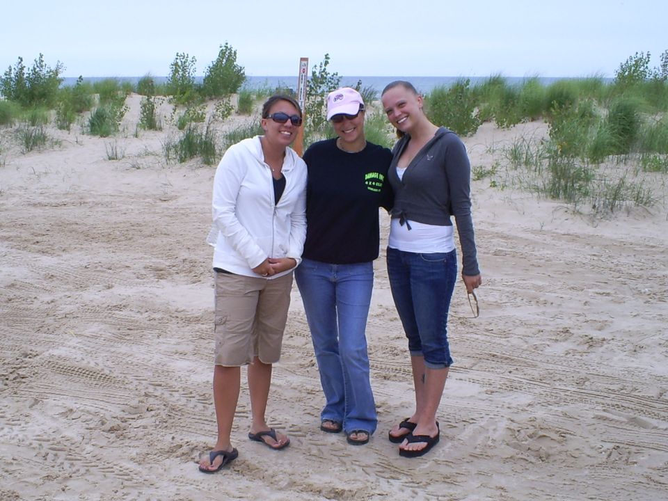 Three women stand on a sandy beach with dunes and water in the background. The woman on the left wears a white zip-up hoodie and tan shorts. The middle woman wears a black t-shirt and blue jeans. The woman on the right wears a grey cardigan and blue ca...